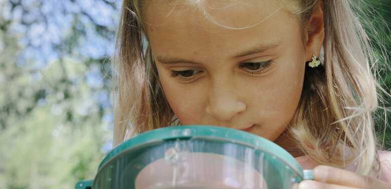Ein kleines Mädchen steht im Wald und beschäftigt sich mit einer Becherlupe.