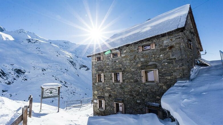 Das Bild zeigt die Essener-Rostocker Hütte im Winter als mehrstöckiges Steingebäude mit schneebedecktem Dach. Vor dem Gebäude liegen hohe Schneemengen, links steht eine Tafel neben einem verschneiten Weg. Im Hintergrund sind schneebedeckte Berghänge zu sehen, während die Sonne knapp über dem Gebirgskamm steht.