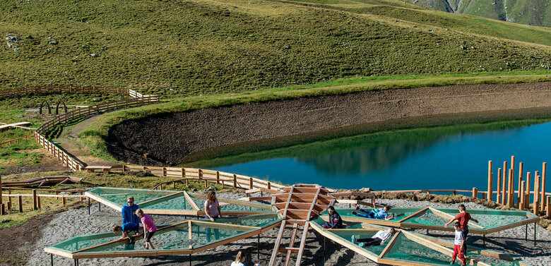 Zu sehen sind mehrere Kinder die auf einem Spielgerät, welches einer überdimensional-großen Libelle ähnlich sieht, beim Wassermythos Ochsenlacke im Skizentrum St. Jakob i. D. spielen. Das sonnige Wetter lässt den sichtbaren Teil des Ufers und die dahinterliegende Bergwelt in einem warmen Licht erstrahlen.