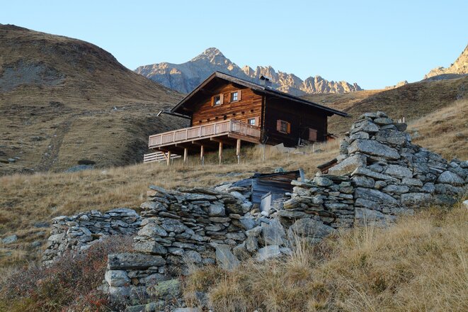 Holzhütte mit Terrasse auf einem Grashang vor felsigem Berggipfel und Steinruinen im Vordergrund.