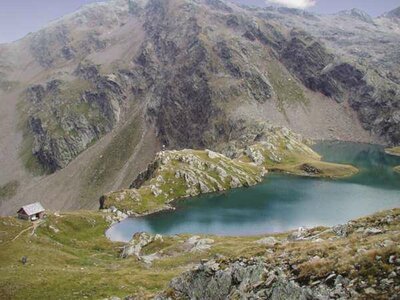 Der Blick von etwas oberhalb auf den Geigensee in Hopfgarten i. D., der seinen Namen seiner geigenförmigen Form zu verdanken hat. Das blaue Wasser hebt sich eindeutig von der grün-grauen, felsigen Umgebung ab. Links vom See steht eine kleine Hütte.