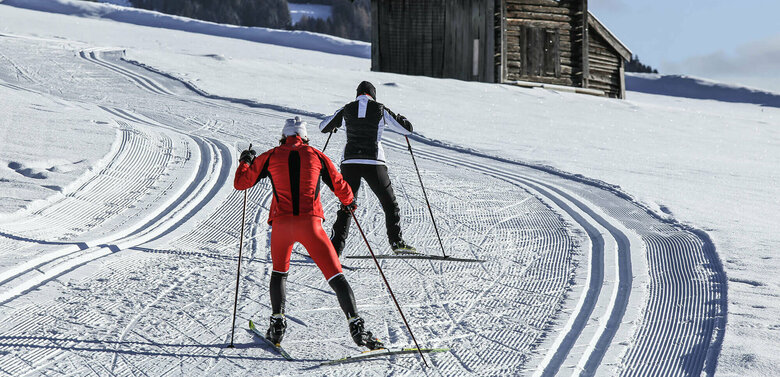 Zwei Langläufer:innen auf der Grenzlandloipe vorbei an einer kleinen Hütte aus Holz