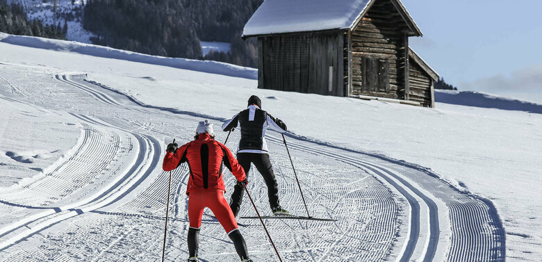 Zwei Langläufer:innen auf der Grenzlandloipe vorbei an einer kleinen Hütte aus Holz