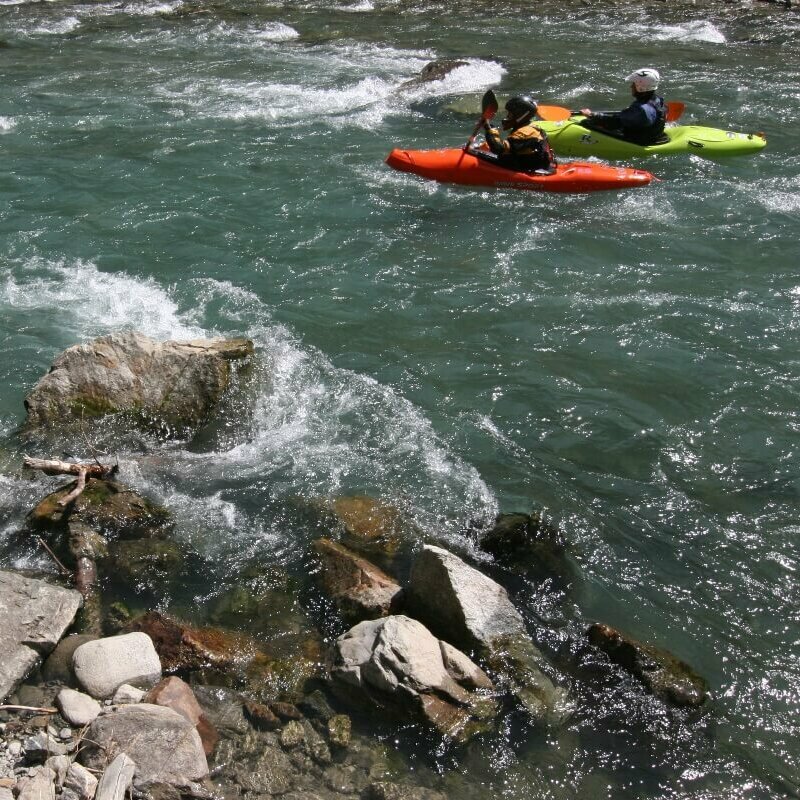 Zu sehen sind drei Kajakfahrer, die in der rauschenden Schwarzach unterwegs sind. Der Wasserspiegel wird von mehreren Felsen durchbrochen.