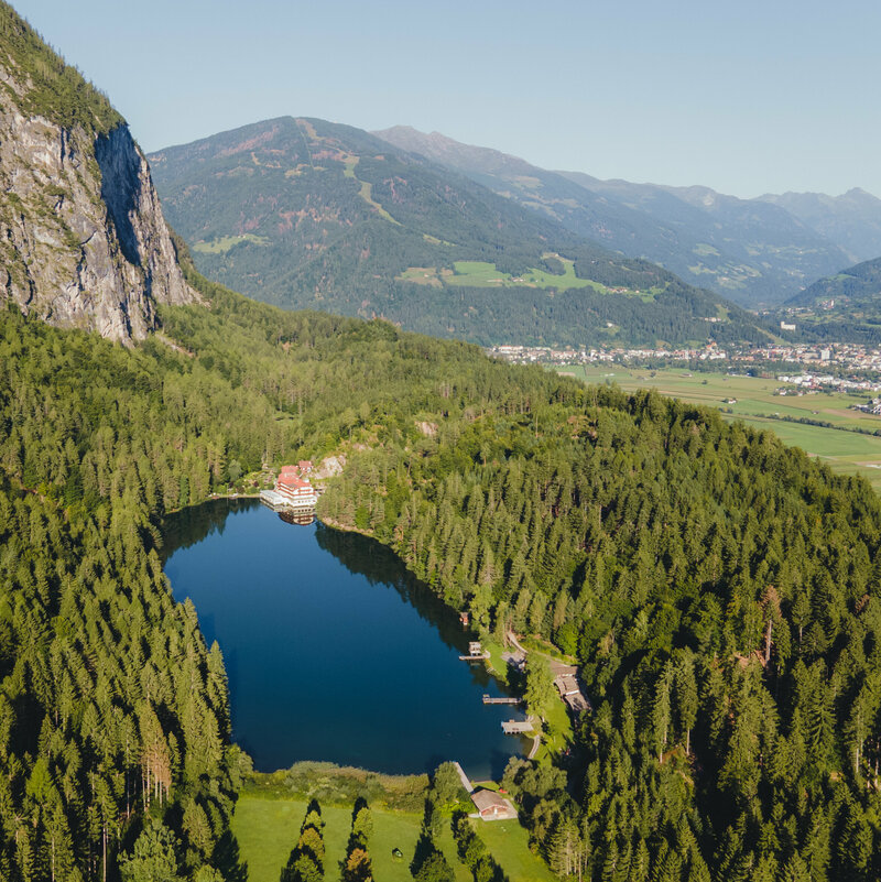 Luftaufnahme vom sanft in den Wald eingebetteten Tristacher See an einem sonnigen Tag, mit Blick auf die Stadt im Hintergrund.