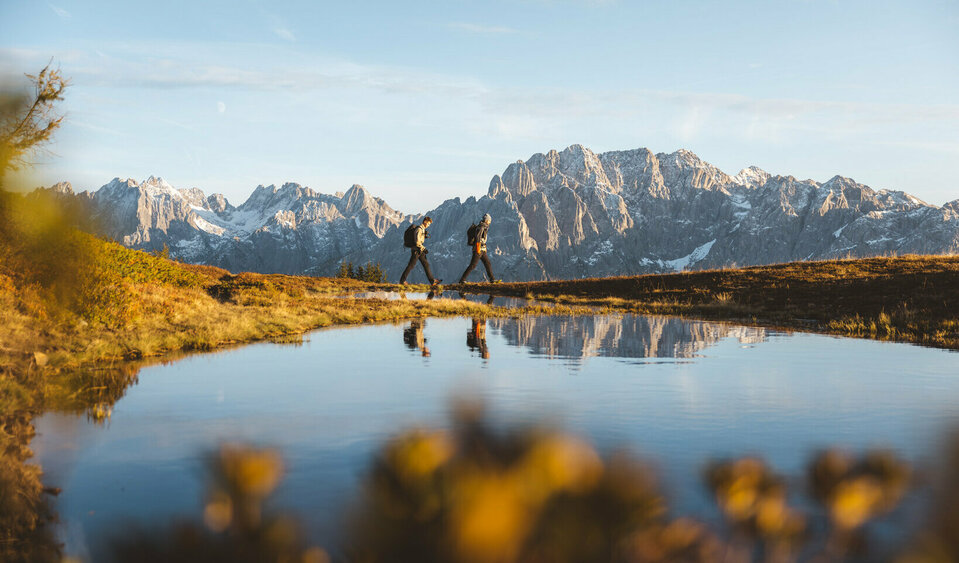 Zwei Personen wandern mit Rucksäcken entlang eines klaren Bergsees. Im Hintergrund erhebt sich eine beeindruckende Alpenkulisse mit schneebedeckten Gipfeln. Das warme Licht der tiefstehenden Sonne beleuchtet die herbstliche Landschaft.