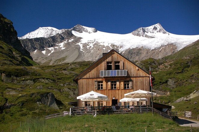 Das Bild zeigt die Johannishütte inmitten einer hochalpinen Berglandschaft. Vor der Hütte befinden sich mehrere Tische mit Sonnenschirmen, während dahinter schroffe Felsberge und vergletscherte Gipfel aufragen. Die Holzfassade der Hütte hebt sich deutlich von den grünen Hängen und dem klaren blauen Himmel ab.