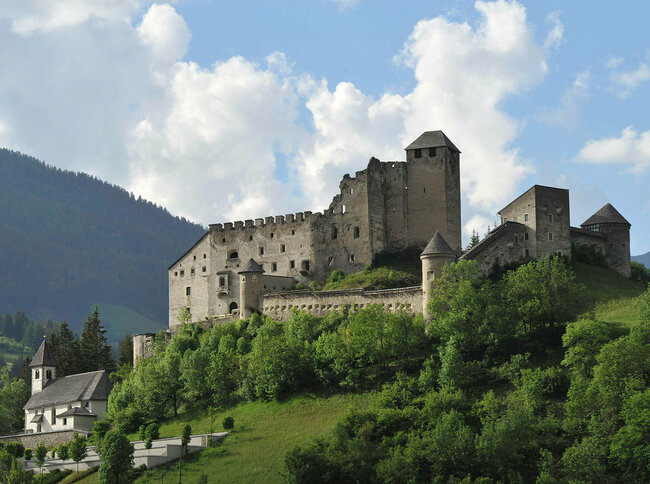 Majestätischer Sommerblick aus dem Tal auf die Burg Heinfels, eingebettet in grüne Laubbäume im satten Sommerkleid