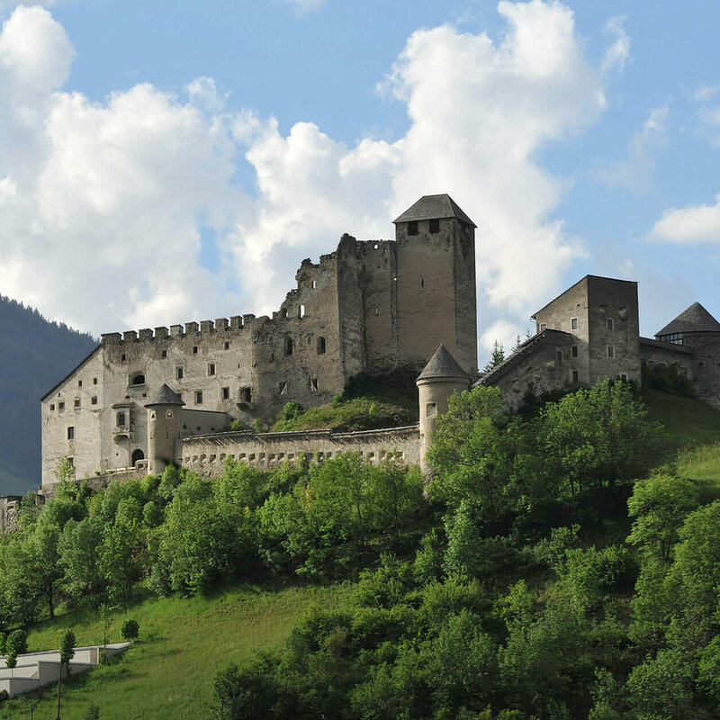 Majestätischer Sommerblick aus dem Tal auf die Burg Heinfels, eingebettet in grüne Laubbäume im satten Sommerkleid