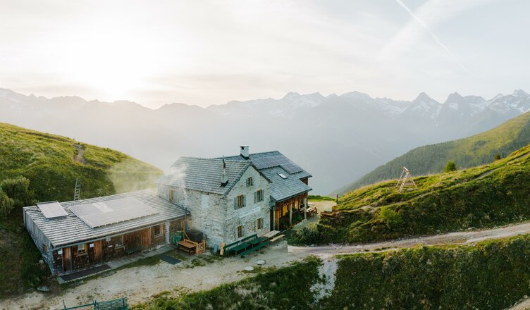 Das Bild zeigt das Kals-Matreier-Törl Haus in hochalpiner Lage auf einem grünen Berghang. Vor der Hütte verläuft ein Fahr- und Wanderweg, während im Hintergrund die Silhouetten zahlreicher Berggipfel zu sehen sind. Das warme Licht und die weite Aussicht unterstreichen die eindrucksvolle Lage der Berghütte.