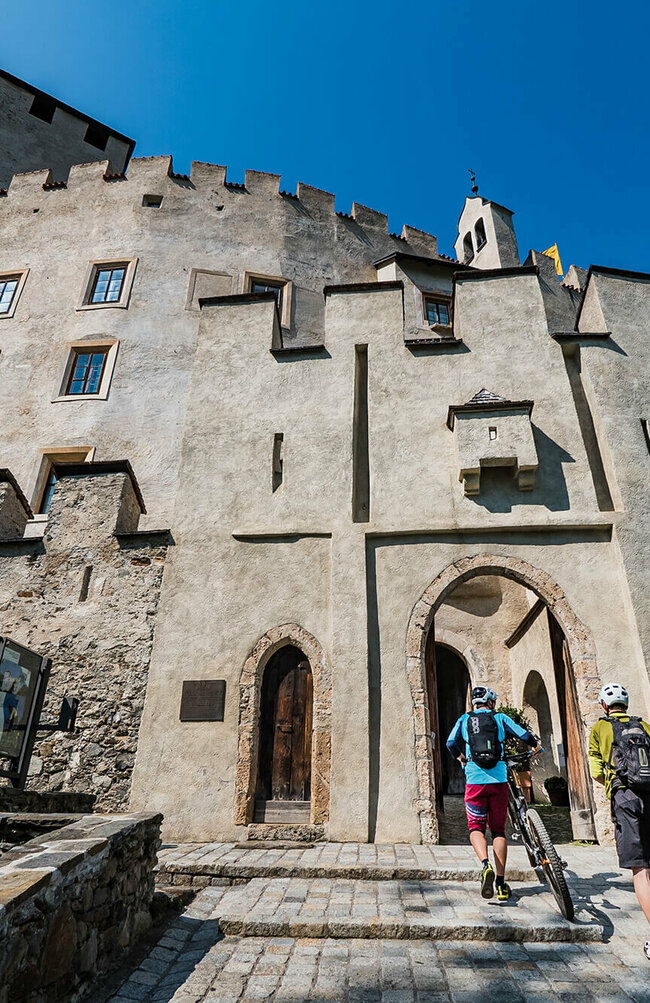 Zwei Radfahrer schieben ihre Fahrräder durch einen steinernen Eingangsbogen in das Schloss Bruck in Lienz.