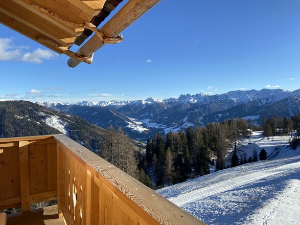 Eine Aussicht von einer Holzterrasse auf schneebedeckte Berge und eine malerische Landschaft. Der klare Himmel und die Sonne verleihen der Szenerie einen besonderen Charme.