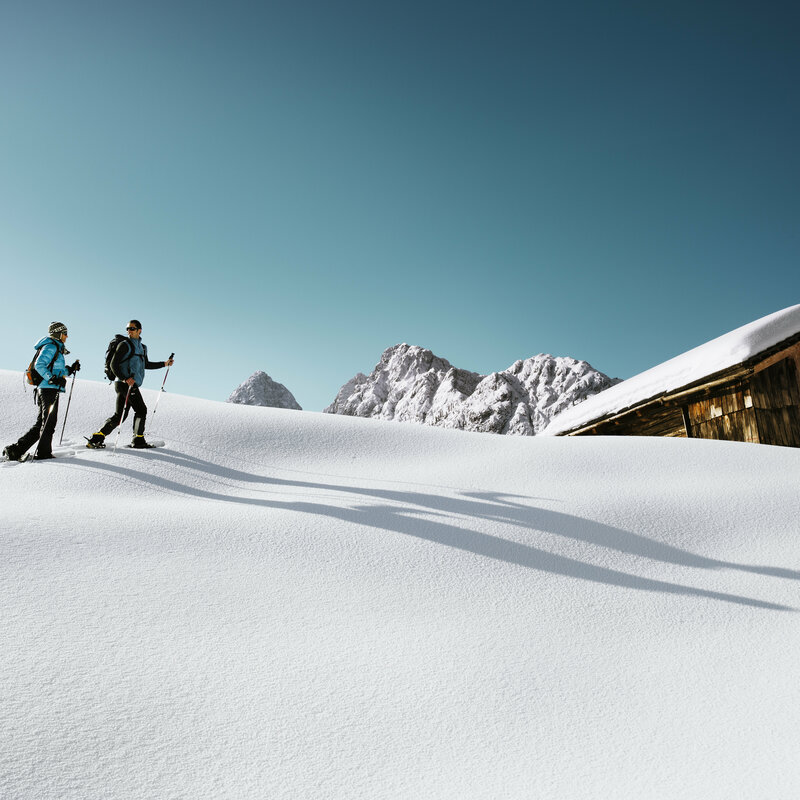 Zwei Personen wandern mit den Schneeschuhen bei schönem Winterwetter durch den Pulverschnee zu einer Hütte.