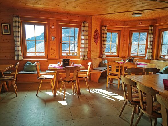 Das Bild zeigt den Gastraum der Badener Hütte mit Wänden, Decke und Möbeln aus hellem Holz. Im Raum stehen mehrere Holztische und Stühle, entlang der Fenster befinden sich gepolsterte Sitzbänke. Durch die zahlreichen Fenster fällt Tageslicht herein und gibt den Blick auf die umliegende Berglandschaft frei.