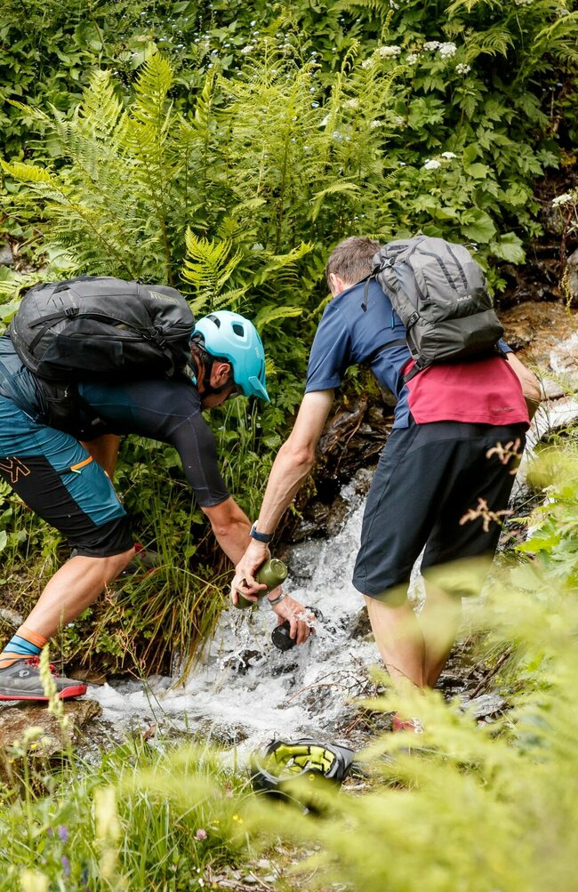 Zwei Männer füllen ihre Trinkflaschen an einem kleinen Bachlauf entlang der Compedalrunde im Kristeinertal.