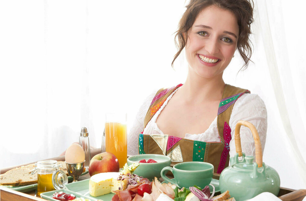 Dame in Dirndl serviert auf einem großen Tableau ein reichhaltiges Osttirol Frühstück mit Speck, Schinken, Käse, Ei, verschiedene Marmeladen und Honig und noch vieles Regionales mehr.