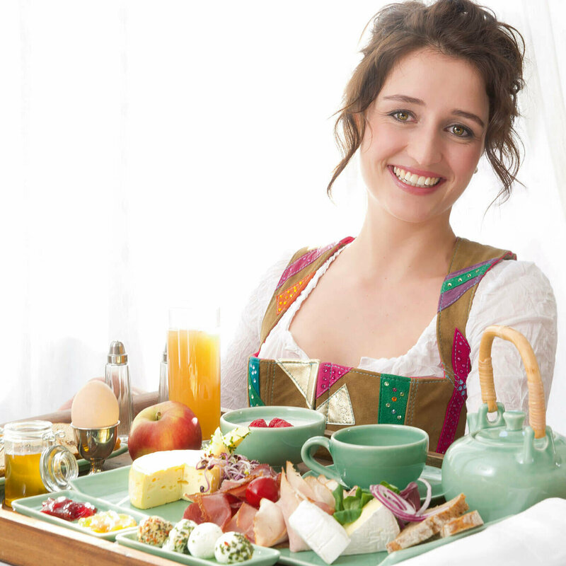 Dame in Dirndl serviert auf einem großen Tableau ein reichhaltiges Osttirol Frühstück mit Speck, Schinken, Käse, Ei, verschiedene Marmeladen und Honig und noch vieles Regionales mehr.