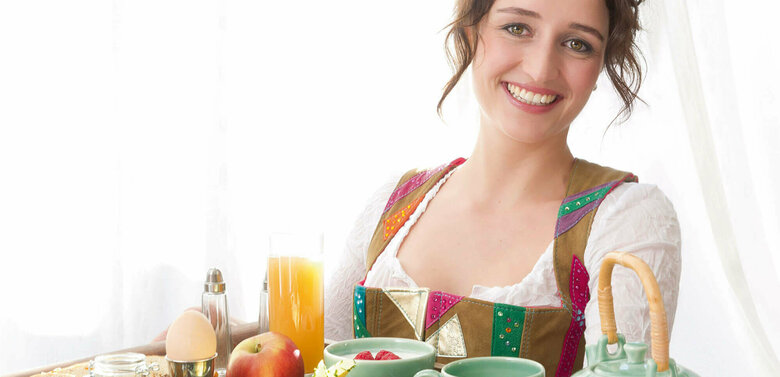 Dame in Dirndl serviert auf einem großen Tableau ein reichhaltiges Osttirol Frühstück mit Speck, Schinken, Käse, Ei, verschiedene Marmeladen und Honig und noch vieles Regionales mehr.