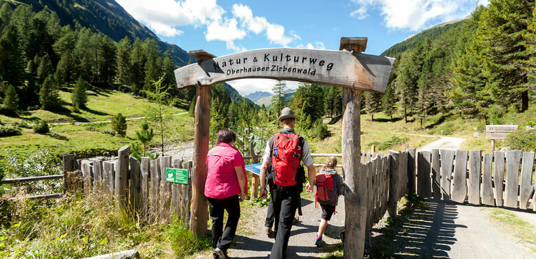 Eine Familie in Wanderkleidung geht durch das aus Holz gebaute Eingangstor am Oberhauser Zirbenwald.