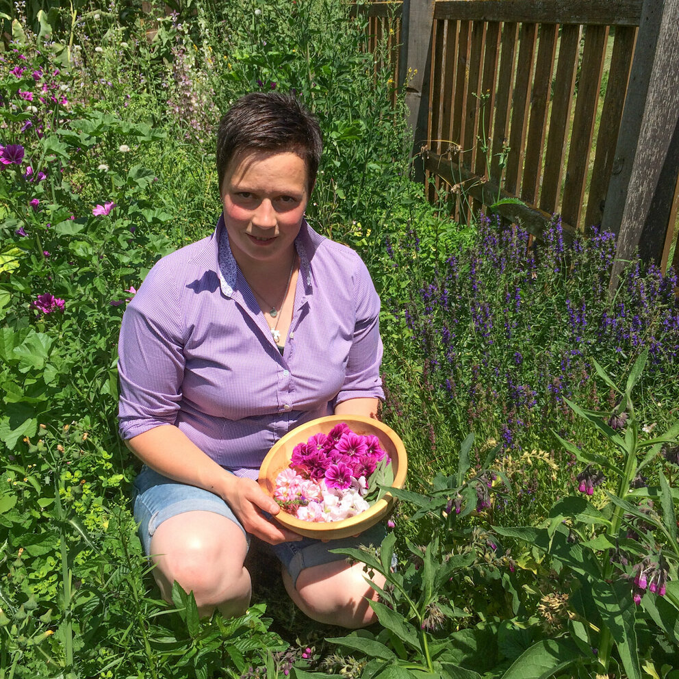 Eine Frau (Viktoria Trager) kniet in einer Blumenwiese und hält einen Holzbehälter mit Blumen gefüllt in der Hand. 