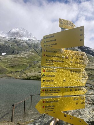 Ein gelber Wegweiser mit zahlreichen Richtungsangaben steht am Ufer eines Bergsees. Im Hintergrund sind grüne Hänge, schneebedeckte Gipfel und die hochalpine Landschaft zu sehen. Die Schilder weisen auf verschiedene Wanderziele in der Umgebung hin.
