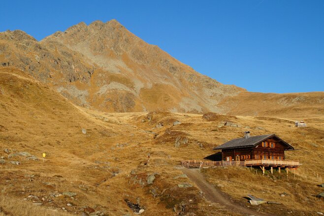 Holzhütte auf einem grasbewachsenen Berghang vor einem felsigen Gipfel unter blauem Himmel.