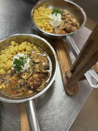 Das Bild zeigt das Islitzer Pfandl in einer kleinen Metallpfanne auf einem Holzbrett. Die Speise besteht aus Spätzle, einer cremigen Sauce sowie Fleisch- und Gemüsestücken und ist mit einem weißen Klecks und frischen Kräutern garniert. Im Hintergrund ist eine weitere Pfanne mit derselben Speise zu sehen.
