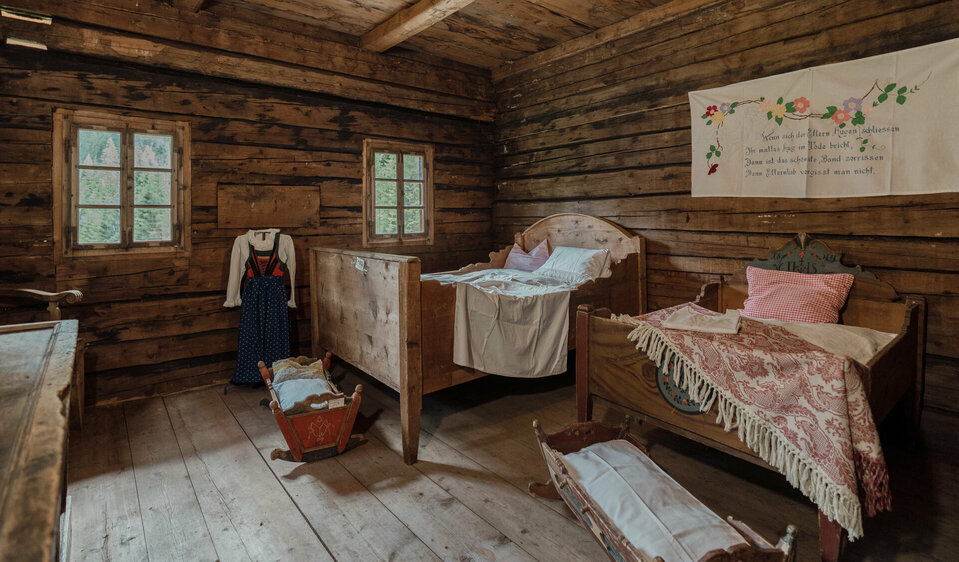 In einem alten Baunerhaus stehen zwei Doppelbetten und zwei Wiegen. Ein traditionelles Trachtengewand wird stolz ausgestellt, während ein kunstvolles Wandtuch die Wand ziert.