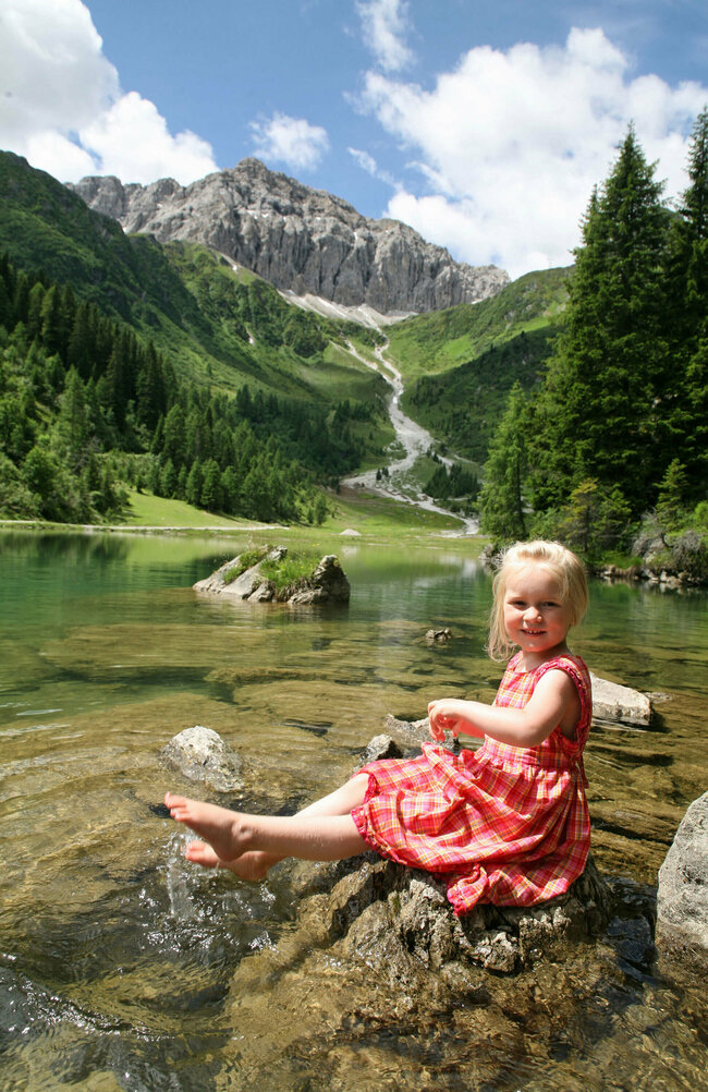 Mädchen in rot-kariertem Sommerkleidchen auf einem Stein am Ufer des Klapfsees sitzend, im Hintergrund rechts mit Nadelbäumen und Felsen begrenztes Seeufer und ganz im Hintergrund das Bergmassiv der Porze aus Kalkgestein. Darüber leicht bewölkter Himmel.
