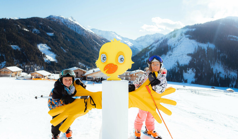 Zwei Kinder stehen im familienfreundlichen Skigebiet Golzentipp in Obertilliach hinter einem gelben Kurvenzeiger in Entendesign mit winterlichem Bergpanorama im Hintergrund.