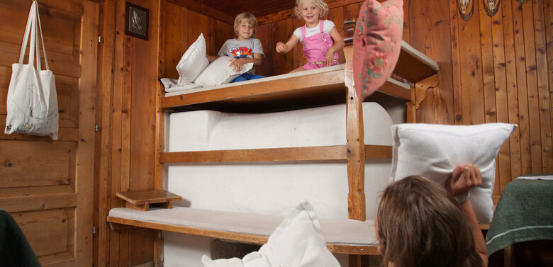 Eine Frau sitzt auf dem Boden in der Stube und hält zwei Kissen in der Hand. Auf dem Ofen sitzen zwei Kinder und werfen Kissen auf die Frau.