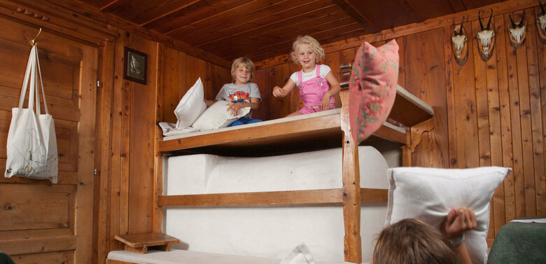 Eine Frau sitzt auf dem Boden in der Stube und hält zwei Kissen in der Hand. Auf dem Ofen sitzen zwei Kinder und werfen Kissen auf die Frau.