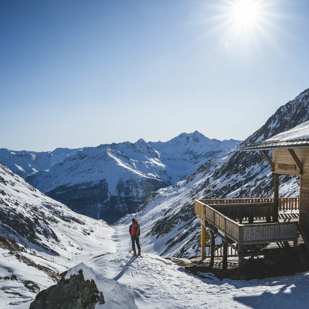 Ein Skitourengeher steht neben der hölzernen Eisseehütte mit Blick talauswärts. Sie Sonne strahlt am blauen Himmel.