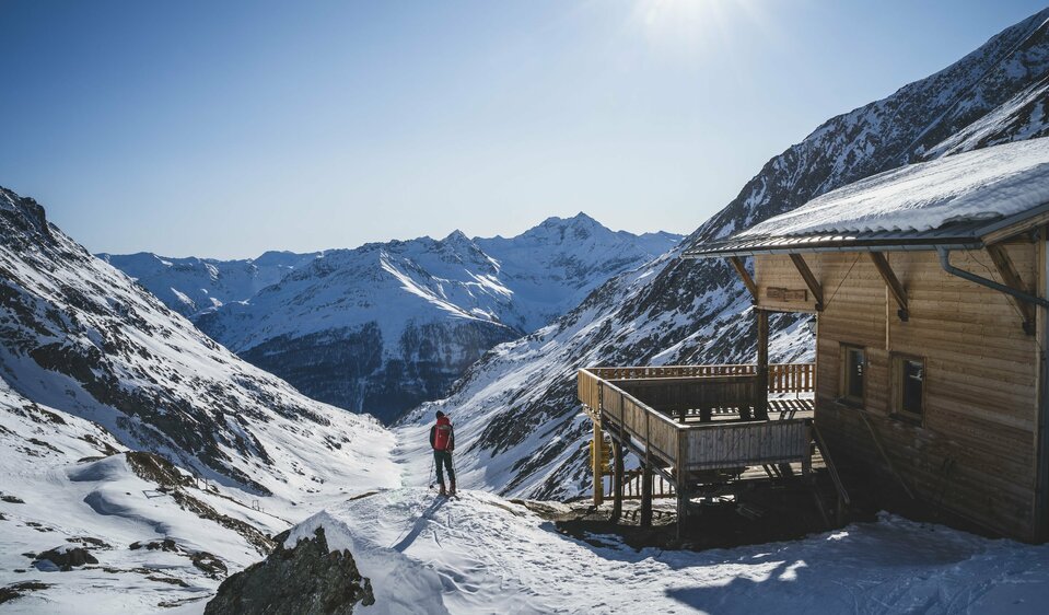 Ein Skitourengeher steht neben der hölzernen Eisseehütte mit Blick talauswärts. Sie Sonne strahlt am blauen Himmel.