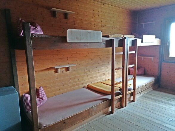 Das Bild zeigt ein Zimmer der Eisseehütte mit Wänden, Decke und Möbeln aus Holz. Im Raum stehen zwei Stockbetten mit Matratzen, Kissen und zusammengelegten Decken. Am Ende des Zimmers befindet sich ein Fenster, durch das Tageslicht hereinfällt.