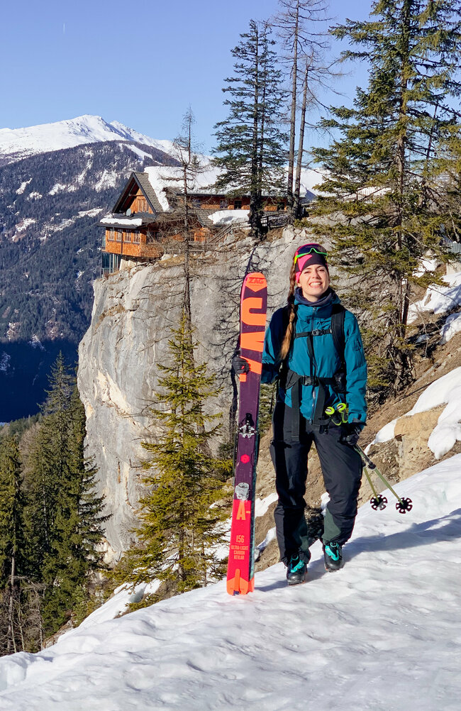 Eine Skitourengeherin steht im Schnee und hält ihre Ski in den Händen, mit der Dolomitenhütte im Hintergrund.