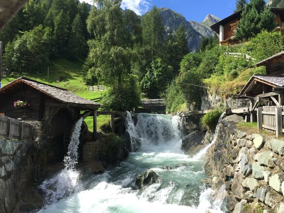 Das Bild zeigt die Islitzer Mühle an einem rauschenden Gebirgsbach. Das Wasser stürzt über mehrere Stufen zwischen Felsen hindurch und fließt unter dem hölzernen Mühlengebäude vorbei. Im Hintergrund sind grüne Wiesen, Bäume und die umliegende Berglandschaft zu sehen.