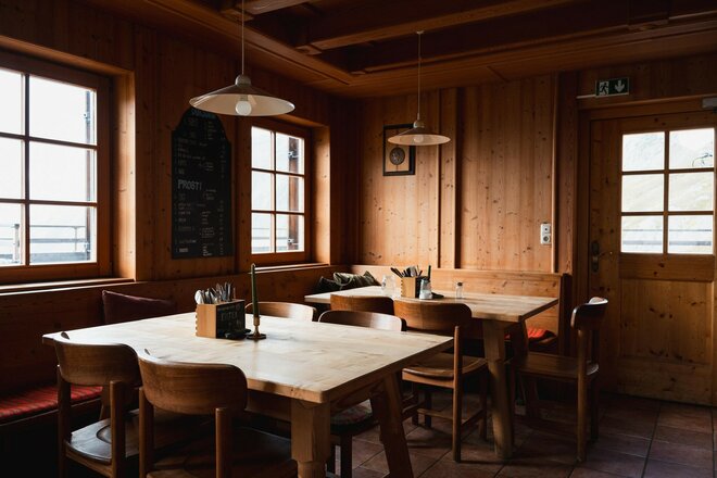 > Die Stube der Sudetendeutschen Hütte ist mit Holz verkleidet und schlicht eingerichtet. Mehrere Holztische und Stühle stehen im Raum, Tageslicht fällt durch die Fenster. Hängelampen und traditionelle Details verleihen dem Gastraum eine gemütliche Atmosphäre.