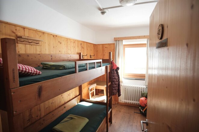 Das Bild zeigt ein Zimmer der Essener-Rostocker Hütte mit zwei Etagenbetten aus Holz. An der Stirnseite befindet sich ein Fenster, vor dem ein Tisch und ein Stuhl stehen. An der linken Wand sind Kleiderhaken mit einer aufgehängten Jacke zu sehen, rechts befindet sich die Zimmertür.