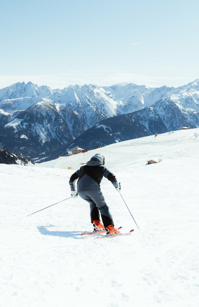 Zwei Skifahrerinnen auf einer flachen Piste oberhalb der Baumgrenze. Im Hintergrund der Taleinschnitt und ein verschneiter Bergkamm, der den Abschluss zur milchig wirkenden Bewölkung an einem Schönwettertag im Winter zeigt.