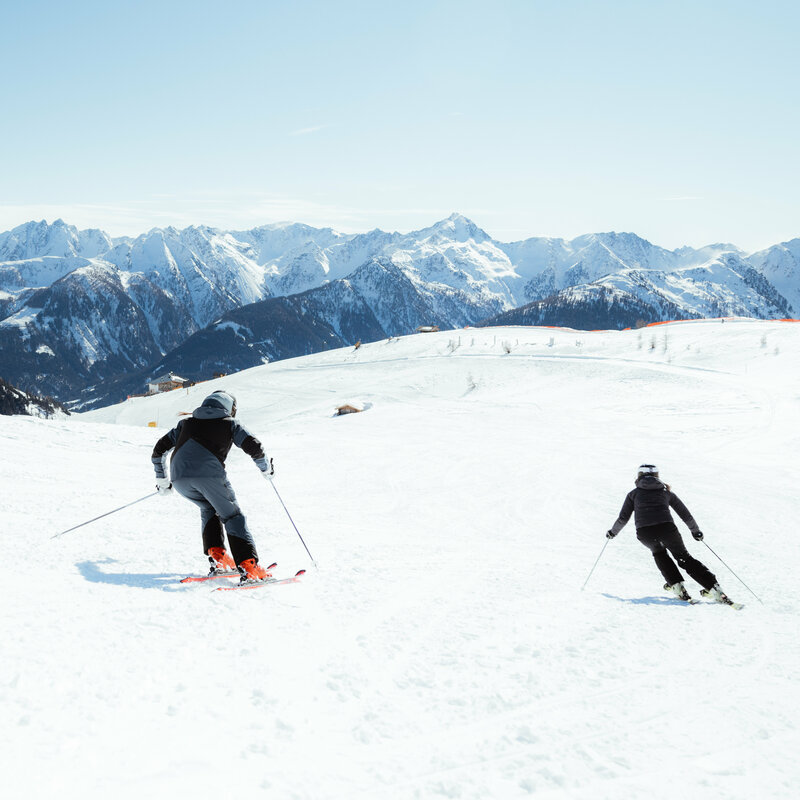 Zwei Skifahrerinnen auf einer flachen Piste oberhalb der Baumgrenze. Im Hintergrund der Taleinschnitt und ein verschneiter Bergkamm, der den Abschluss zur milchig wirkenden Bewölkung an einem Schönwettertag im Winter zeigt.