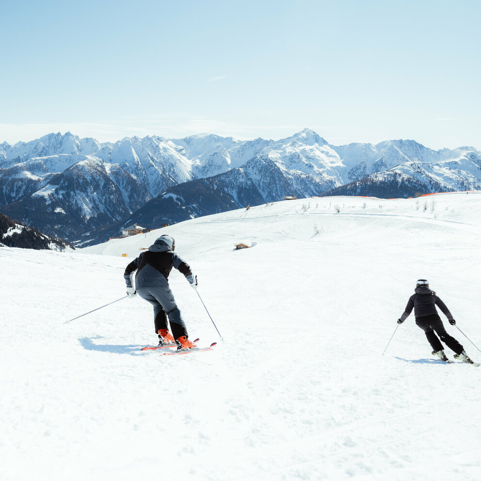 Zwei Skifahrerinnen auf einer flachen Piste oberhalb der Baumgrenze. Im Hintergrund der Taleinschnitt und ein verschneiter Bergkamm, der den Abschluss zur milchig wirkenden Bewölkung an einem Schönwettertag im Winter zeigt.