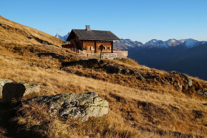 Holzhütte an einem Grashang vor schneebedeckten Berggipfeln unter blauem Himmel.
