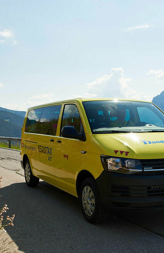 Das gelbe Autosammeltaxi Osttirol führt über eine Straße, daneben befindet sich eine Blumenwiese. Im Hintergrund sind Berge.