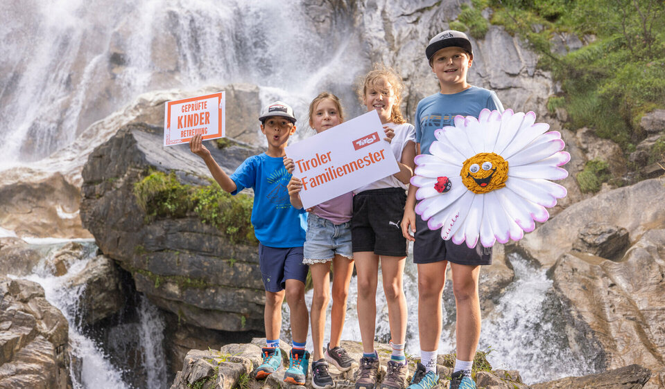Vier Kinder stehen mit Gisela Gänseblümchen vor einem Wasserfall, während zwei von ihnen die Schilder der Tiroler Familiennester in die Höhe halten.