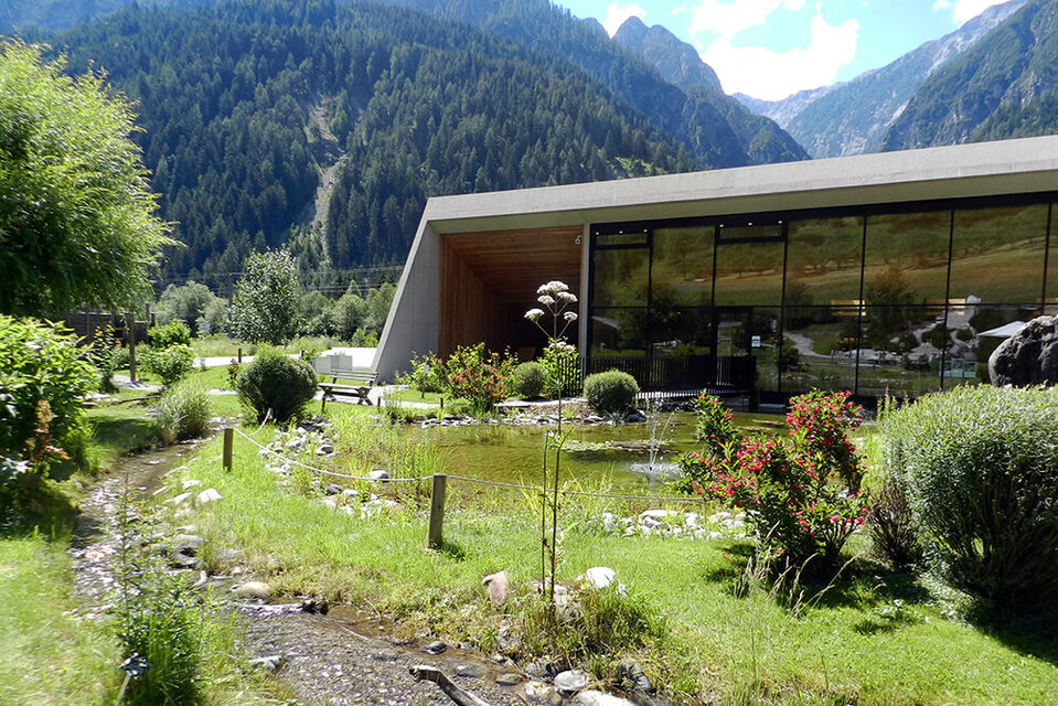 Ein sanft plätschernder Bach schlängelt sich durch den Wohlfühlgarten Vitalpinum, während im Hintergrund der einladende Eingangsbereich mit dem Shop sichtbar ist.