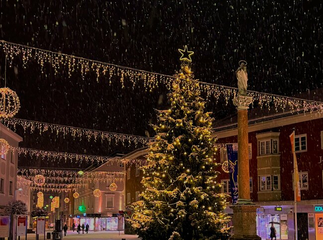 Blick vom Johannesplatz in Richtung Hauptplatz mit einem lichterverzierten Tannenbaum und dichtem Schneefall.