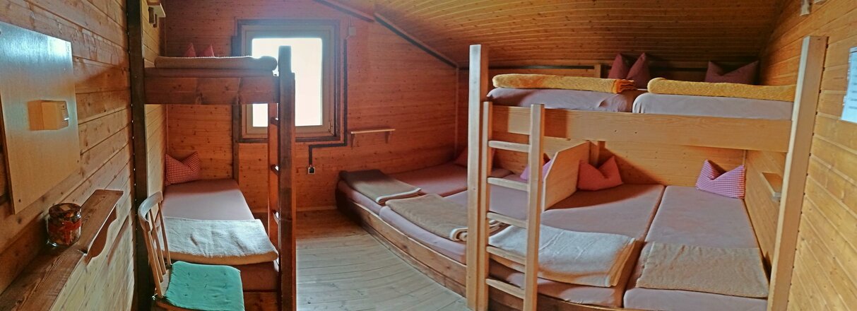 Das Bild zeigt ein Zimmer der Eisseehütte mit Wänden, Decke und Möbeln aus Holz. Im Raum befinden sich zwei Stockbetten sowie ein Doppelbett, die mit Matratzen, Kissen und Decken ausgestattet sind. Am Ende des Zimmers befindet sich ein Fenster, durch das Tageslicht hereinfällt.