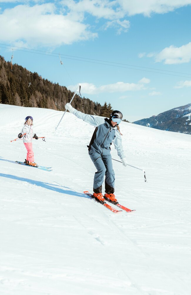 Ein Erwachsener fährt mit einem Kind auf einer flachen Piste im Skigebiet Golzentipp in Obertilliach bei herrlichem Winterwetter.
