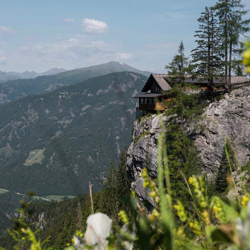 Die Dolomittenhütte steht auf einem Plateau vor einem Abgrund in sommerlicher Landschaft.